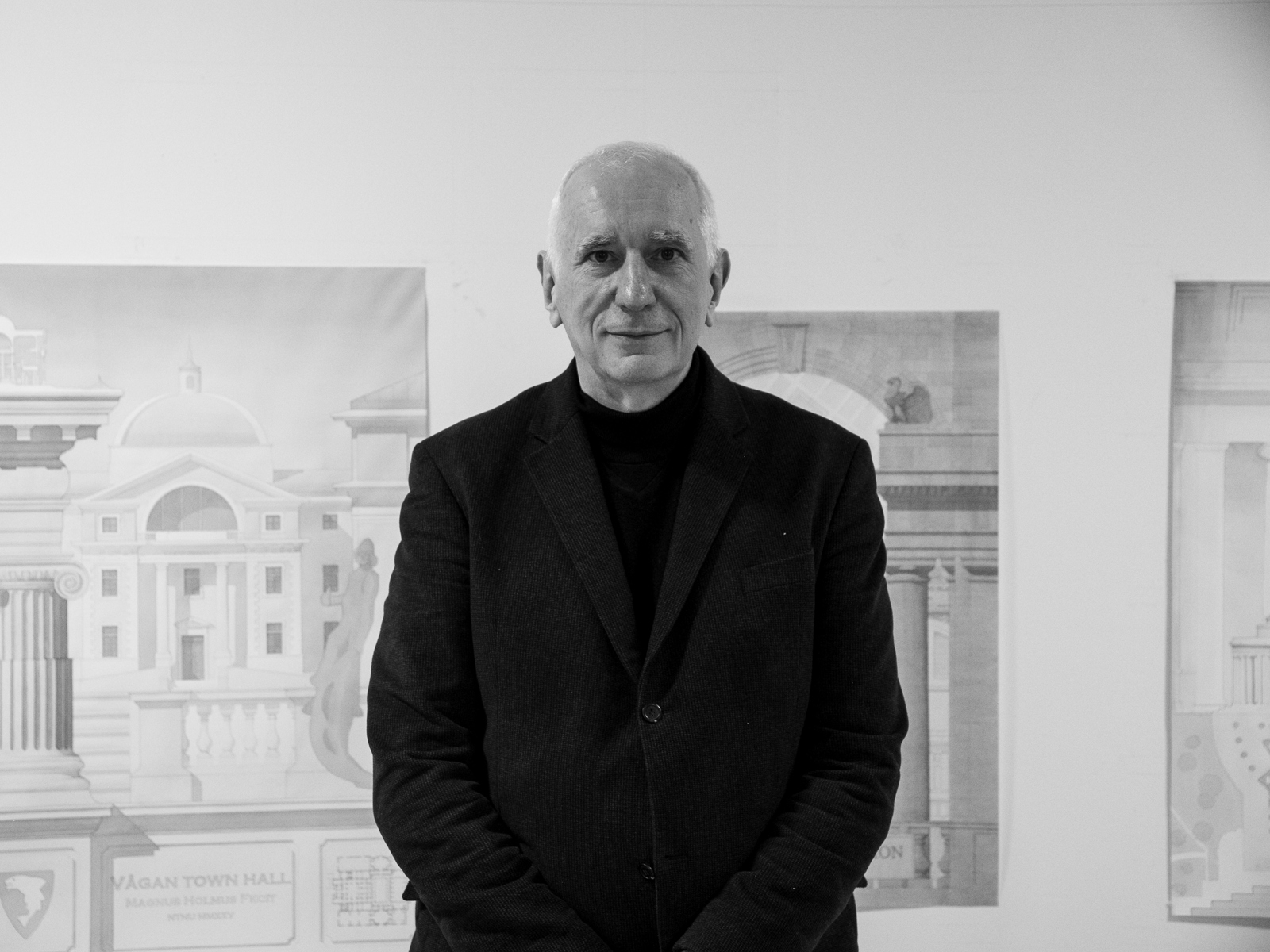 Svart hvitt portrett foto av Branko Mitrovic fremfor flere akvareller av bygninger.