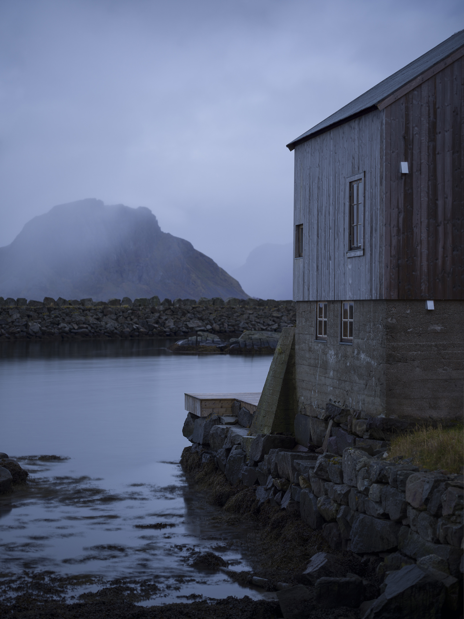 Foto av et grånet bygg med høy grunnmur i betong i havgapet, fjell i bakgrunnen og tåke. 