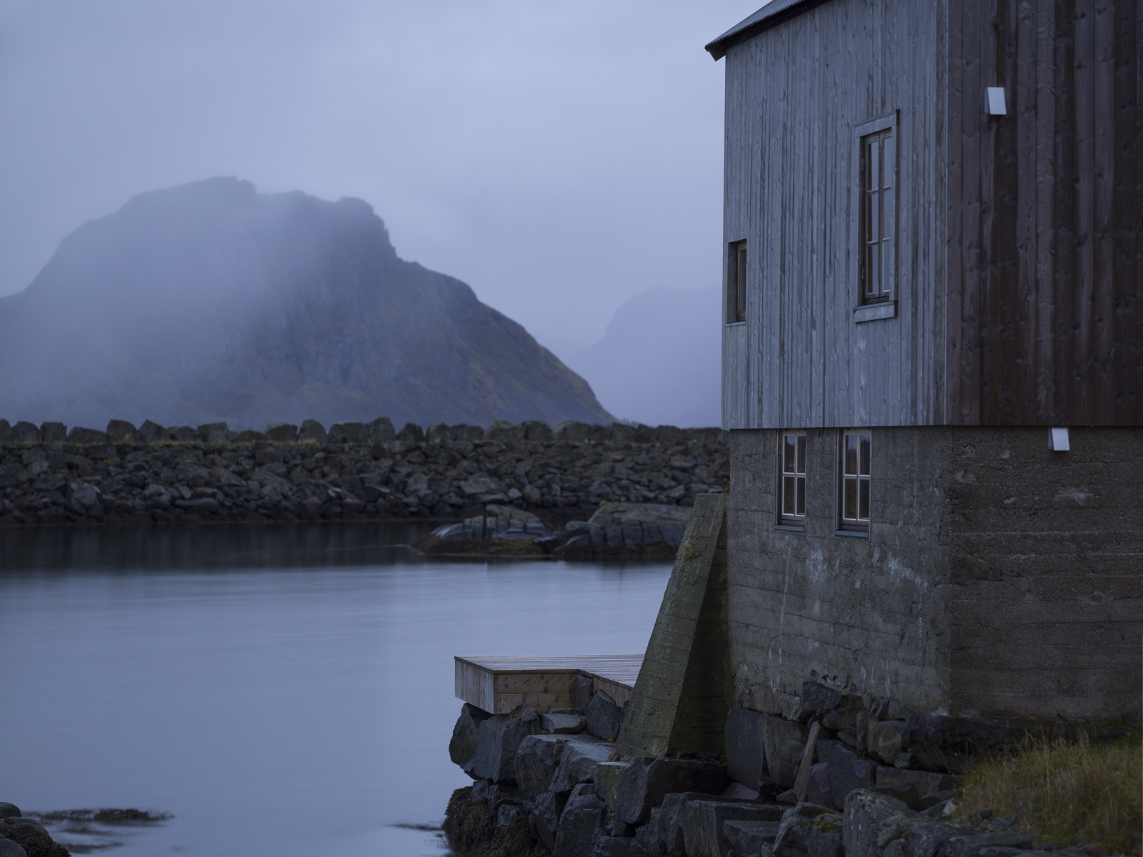 Foto av et grånet bygg med høy grunnmur i betong i havgapet, fjell i bakgrunnen og tåke. 