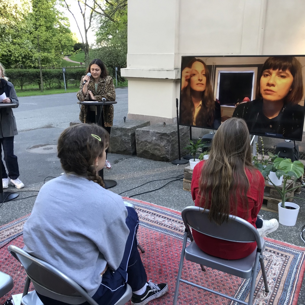 TV satt opp ute, 2 damer som står å snakker i hver sin mikrofon fremfor hvert sitt runde bord, klappestoler med publikum på et teppe. Foto.