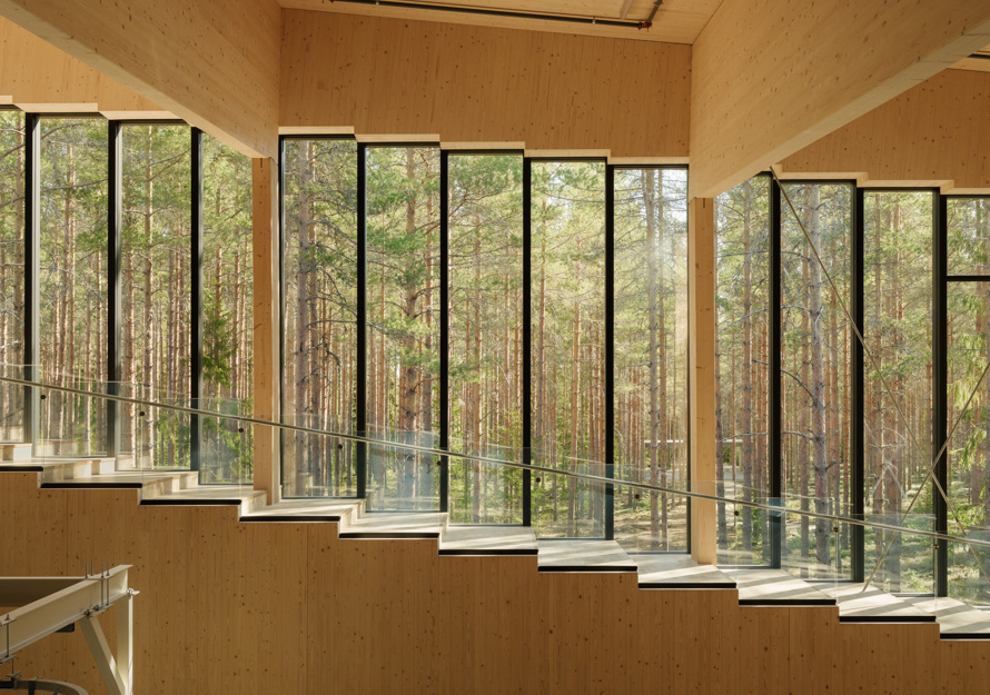 Interiør i treverk, smale vertikale vinduer dekker veggen og man kan se skogen utenfor. Foto.