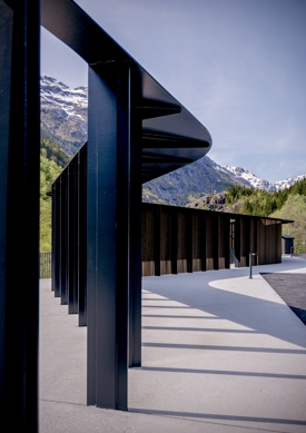 Mørk metallstruktur slynger seg over en parkeringsplass med fjell i bakgrunnen. Foto.