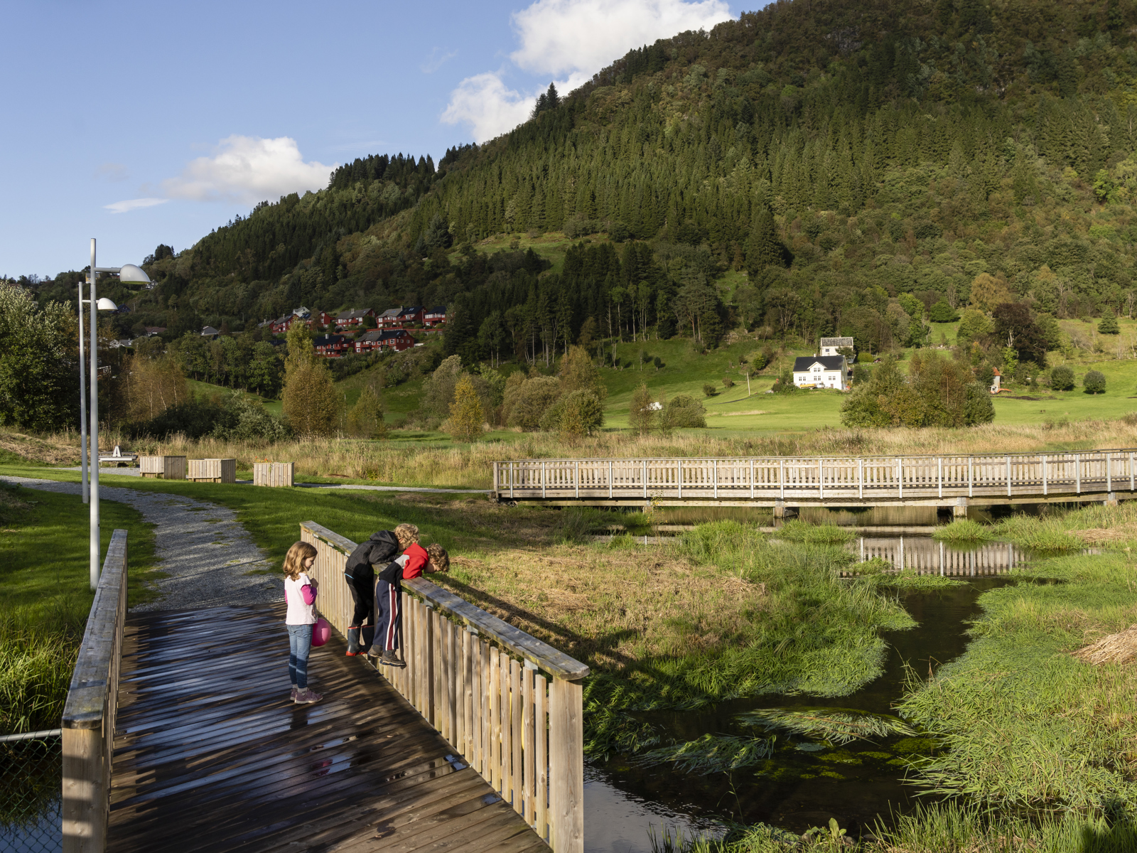 Lekeplass med trestolper og barn som leker, åskam i bakgrunnen. Foto.