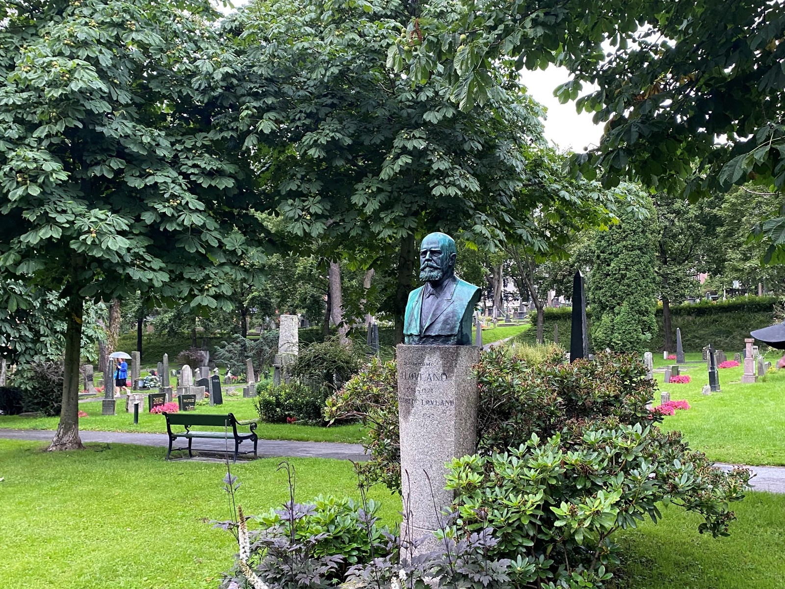 Byste av mann i staudebed på gravlund. Foto.
