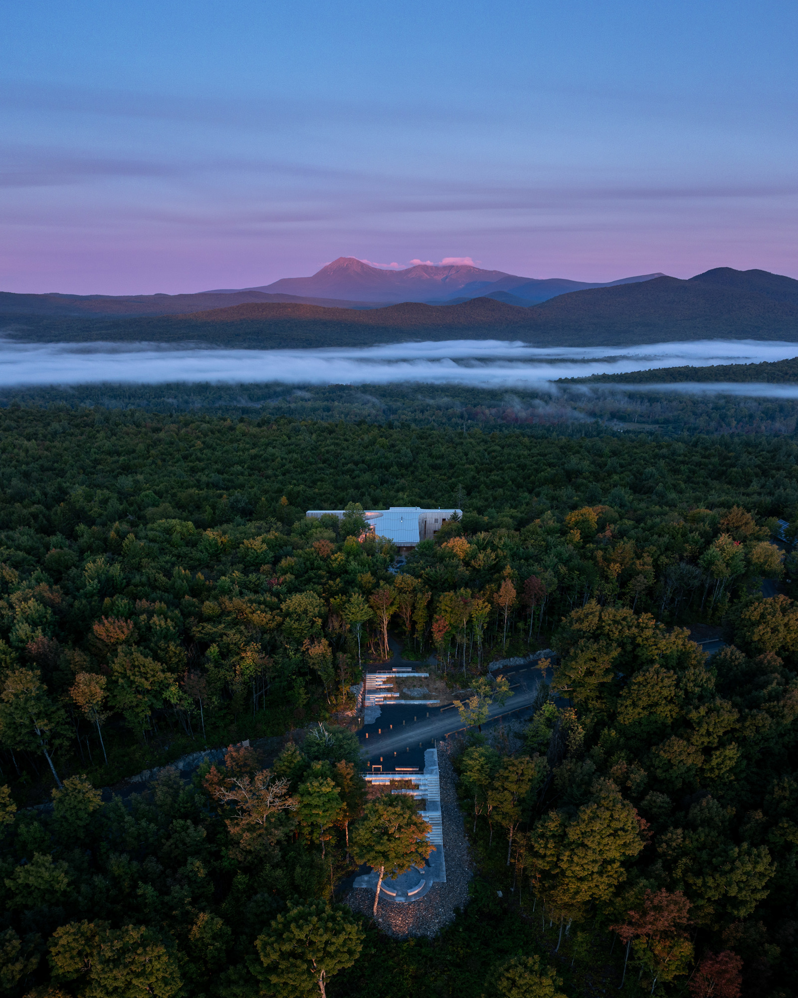 Drone foto av et bygg som ligger helt alene i en stor skog, fjell i bakgrunnen, rosa lys fra sola som akkurat har gått ned.