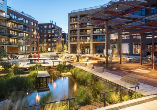 Foto av Vollebekk torg med variert bygulv, åpent takoverbygg og ulike sitteplasser, kveldslys.