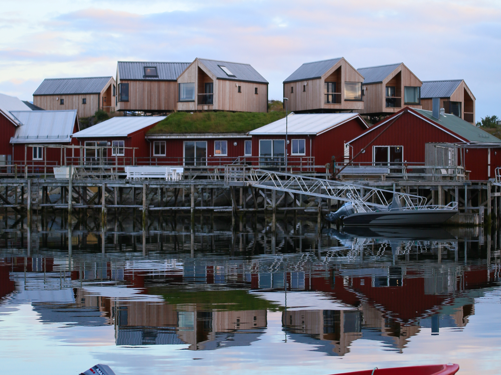 Foto av brygge med små røde hytter og hytter i lyst treverk bak, vann med en rød båt i forgrunnen. 