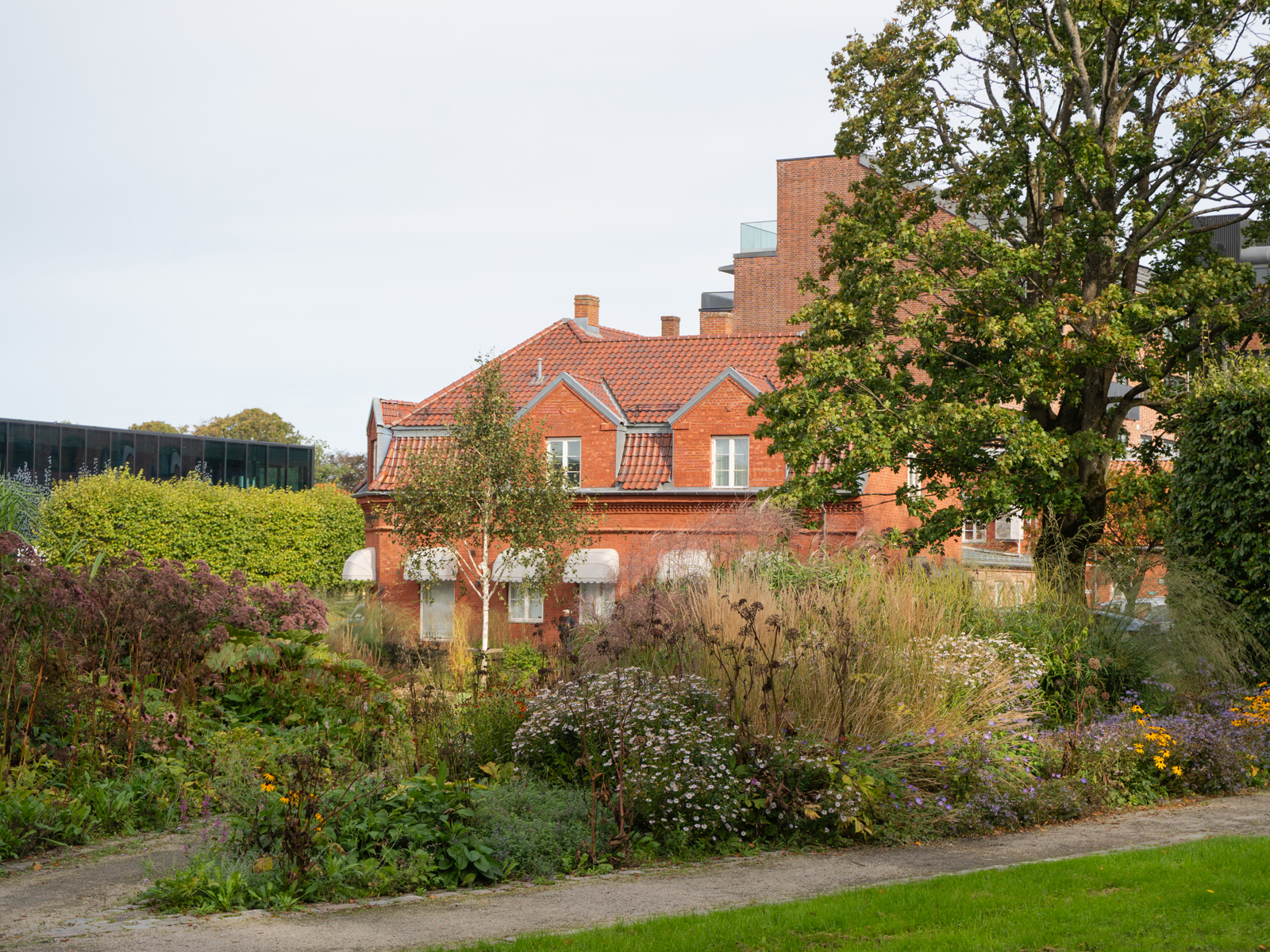 Foto av et stort plantefelt med mye ulik vegetasjon, innrammet av grussti, et rødt teglhus bakenfor. 
