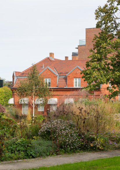 Foto av et stort plantefelt med mye ulik vegetasjon, innrammet av grussti, et rødt teglhus bakenfor. 