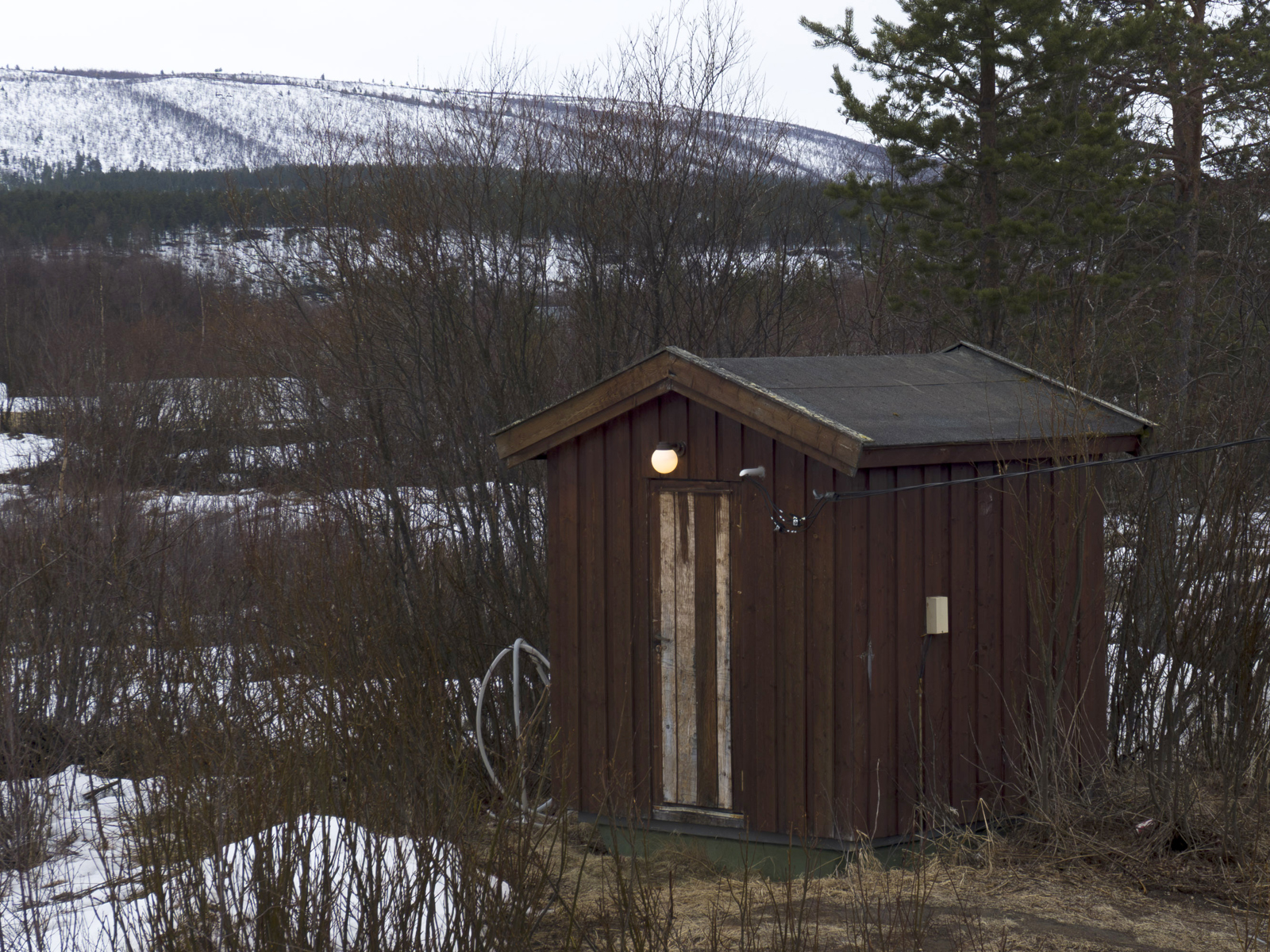 Foto av en liten hytte i naturen, litt snø rundt om. 