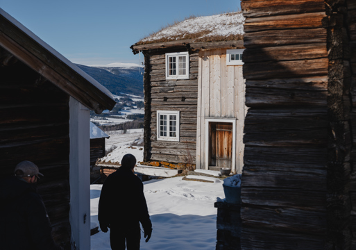 Foto av en mann som går mellom eldre hus på et gårdstun i vinterlandskap.