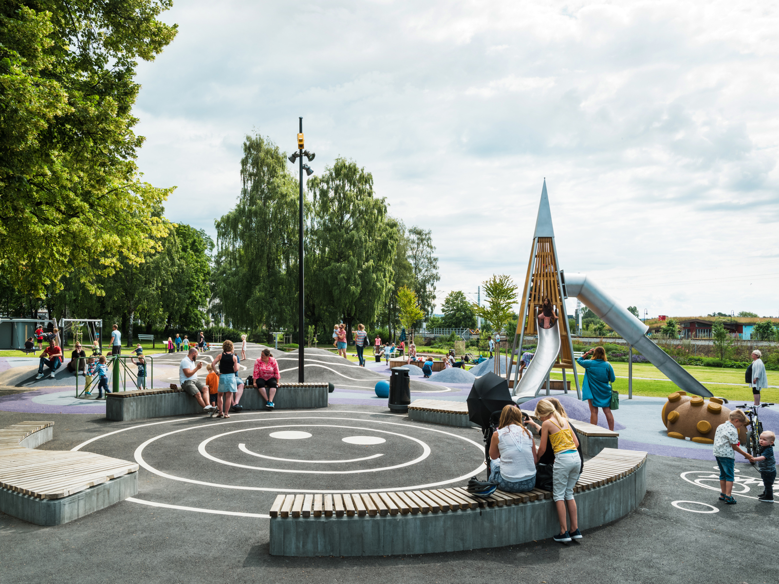 Foto av lekeplassen i Strandgateparken, benker og mange ulike lekeapparater, barn leker og voksne passer på.
