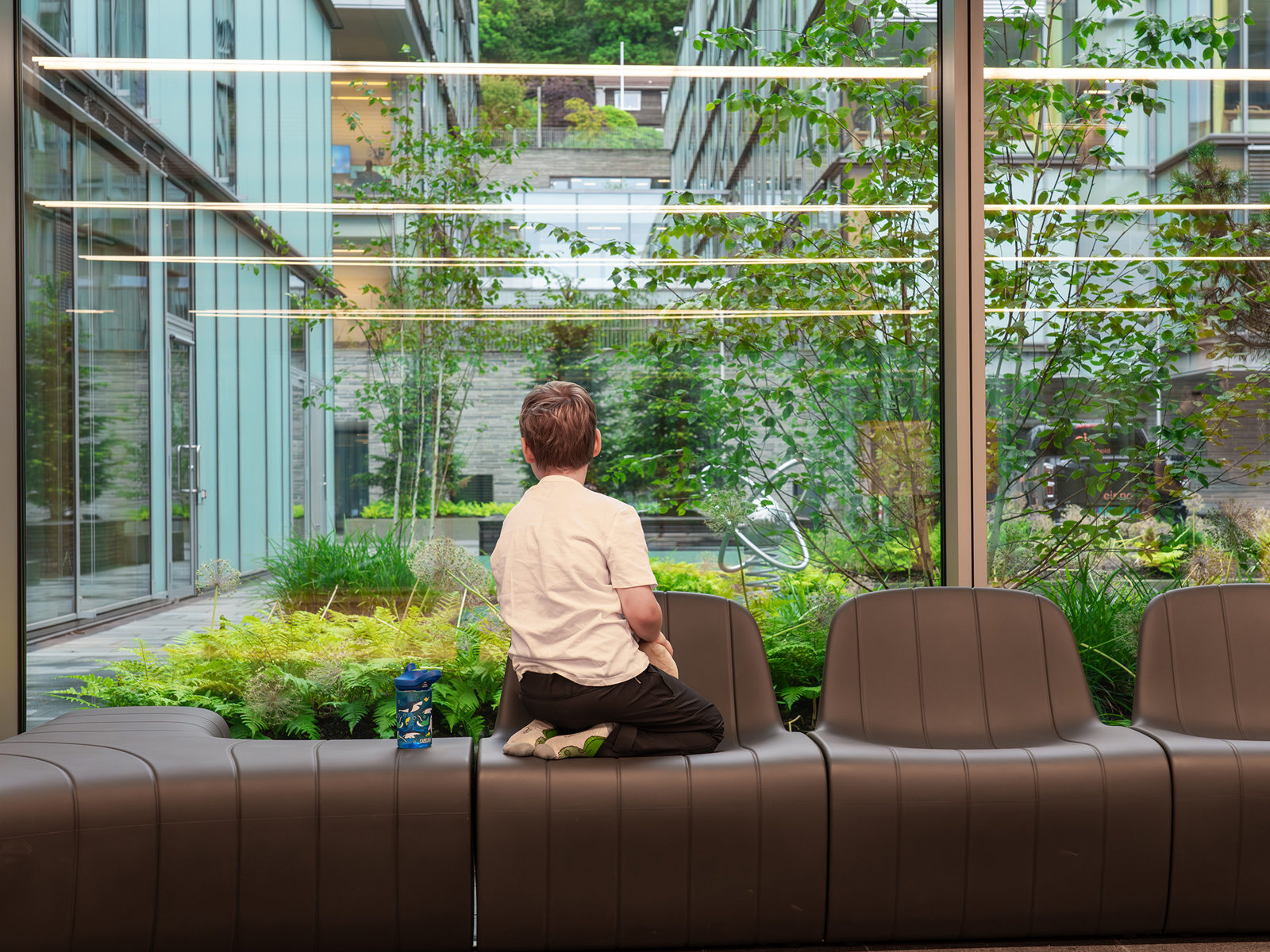 Interiør foto av en gutt som sitter på en brun sofa og titter ut gjennom store vindusflater til et grønt uterom.