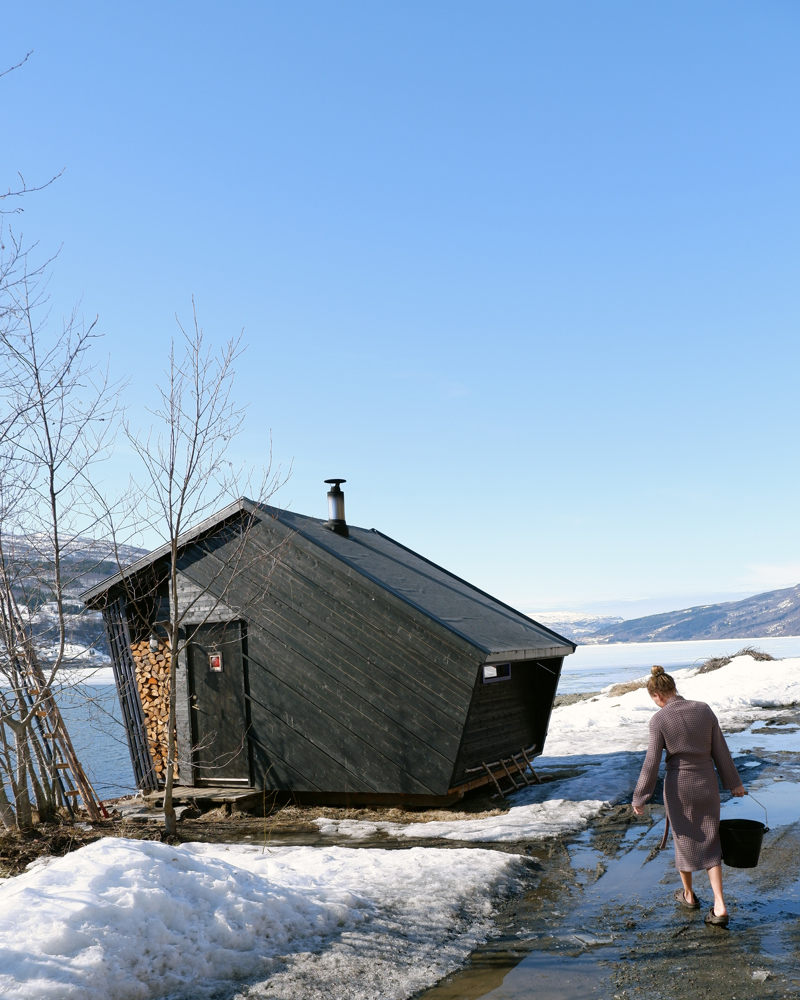 Foto av svart malt sauna i vinterlandskap med vann og fjell i bakgrunnen, en dame bærer en bøtte vann til saunaen. 