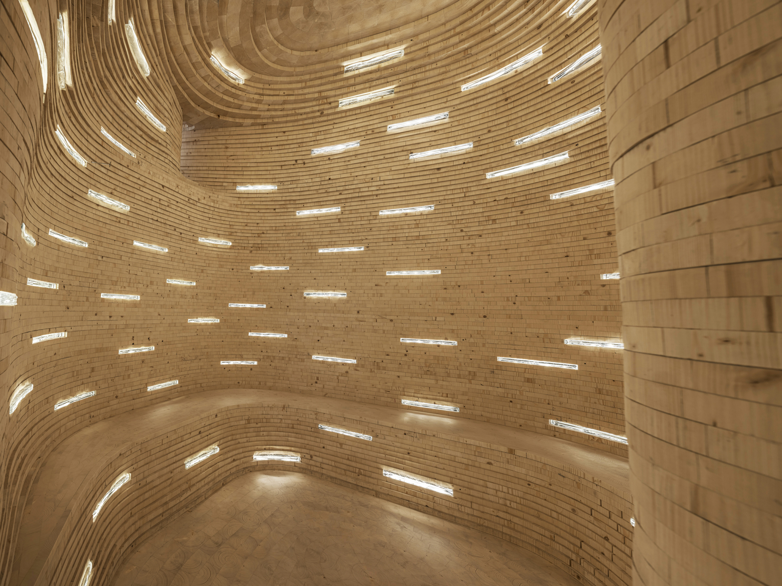 Innsiden av et organisk krummet rom bygget opp av stablet tre lameller, med horisontale smale åpninger som slipper inn lys. Foto.