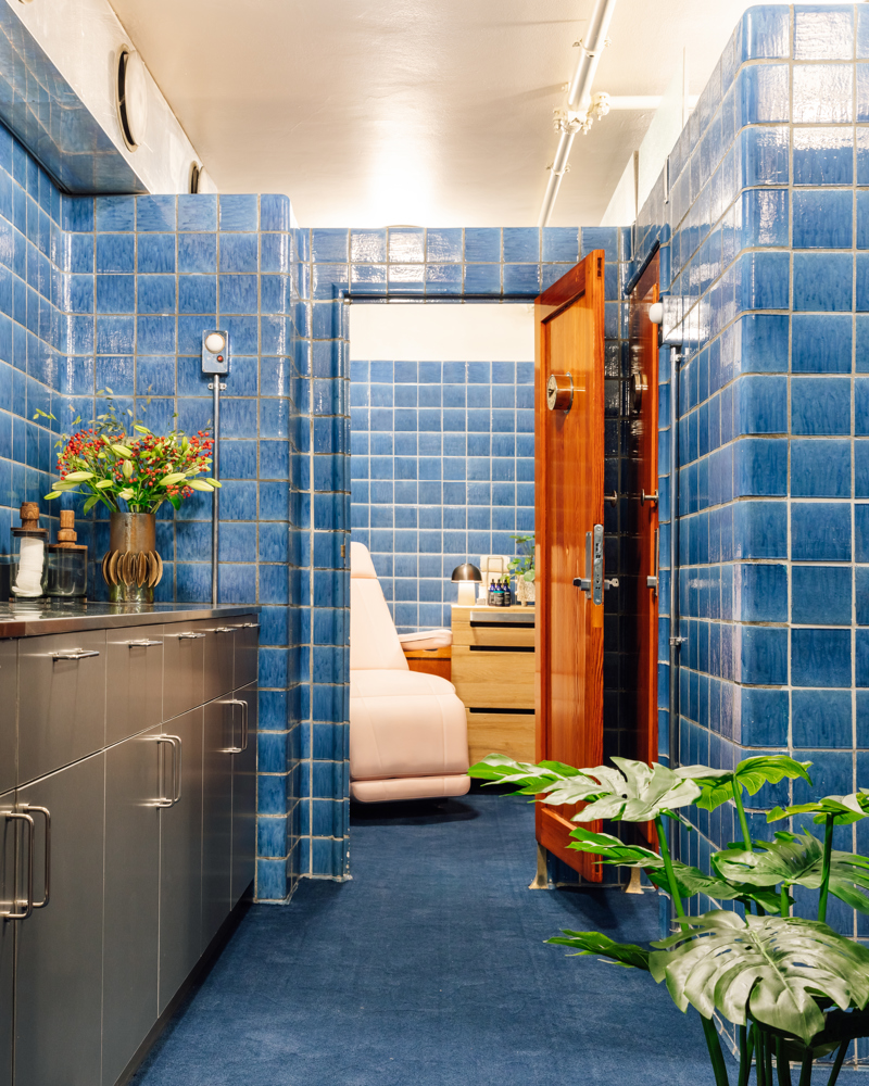 Interiør, rom med blå fliser på vegg og gulv, stålbenk og åpen dør til en stol. Foto.