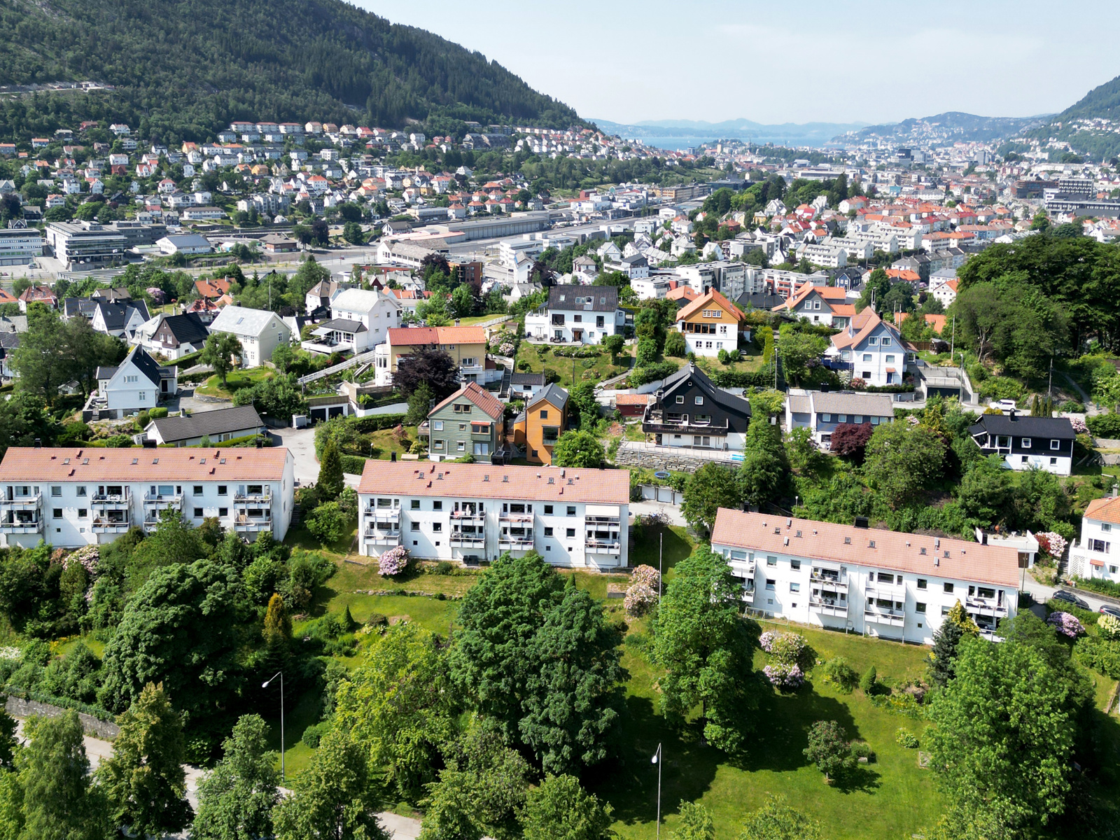 Oversiktsfoto av nabolag, med elv, trær og grønt i front, by, åser og hav i bakgrunnen. 