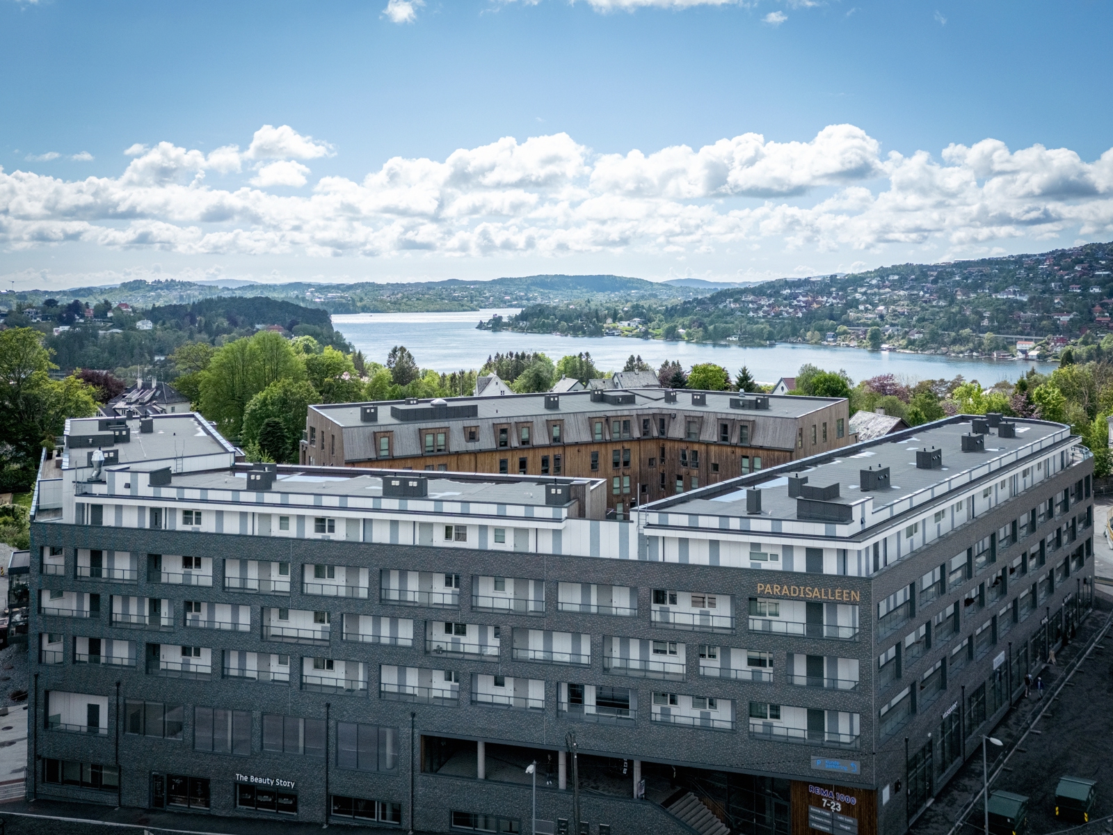Oversiktsbilde av bolig kompleks, grå fasade, vann og åser i bakgrunnen. Foto.