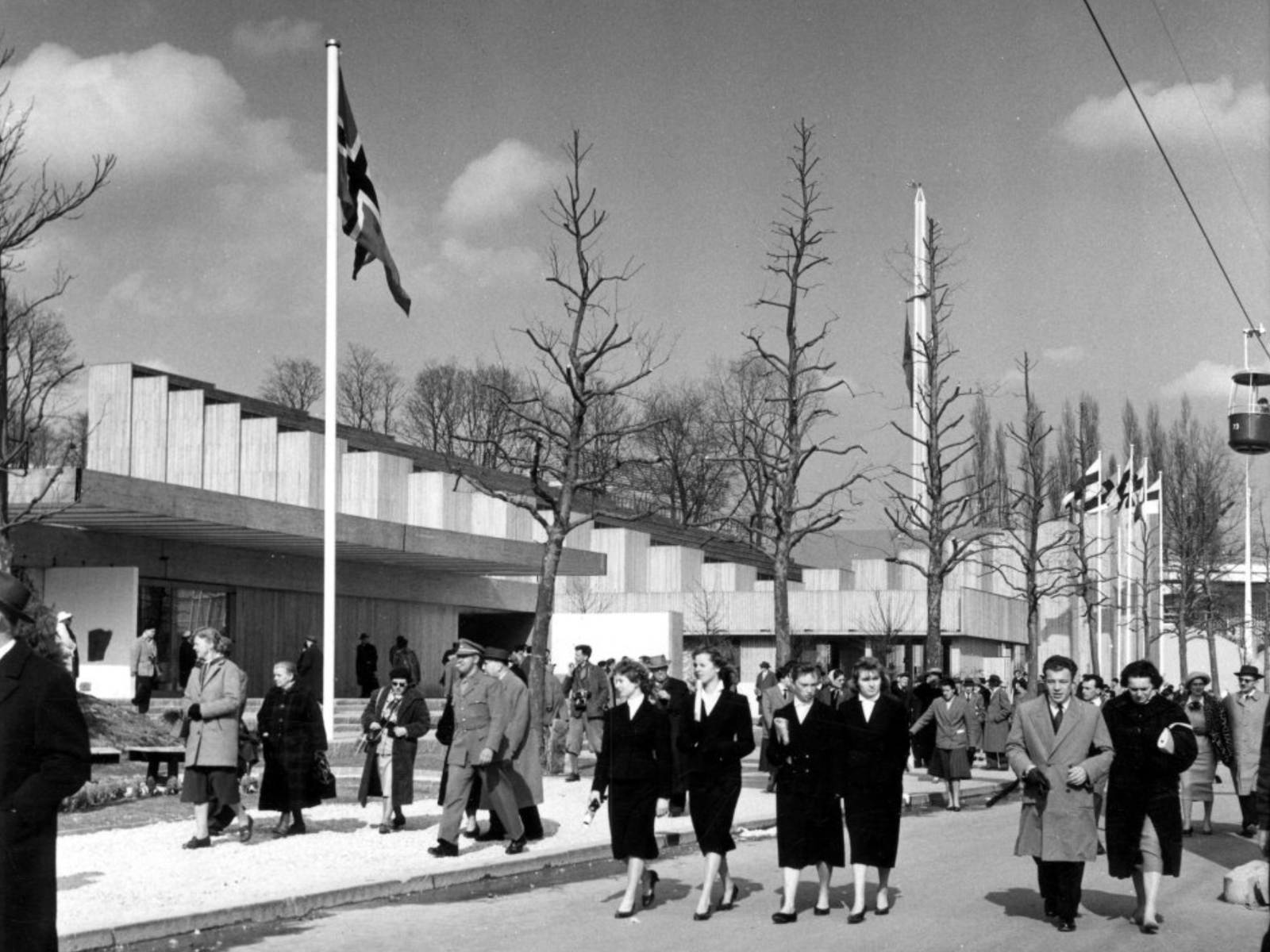 Svart hvitt foto av Expo58 i Brussel, pent kledde mennesker, norsk flagg, paviljong i bakgrunnen.