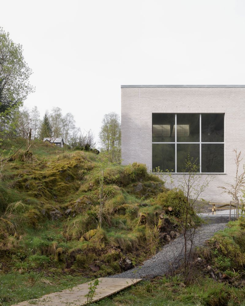 Foto av hvit teglsteinsbygg med store vindusflater, mosegrodd landskap med sti fremfor.