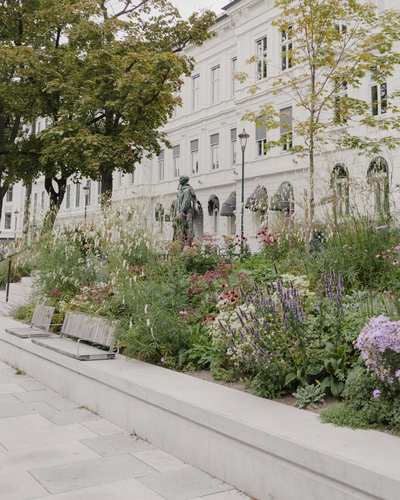 Foto av opphøyde plantefelt ved fortau, med benk langs kanten, statue og bygg i bakgrunnen.