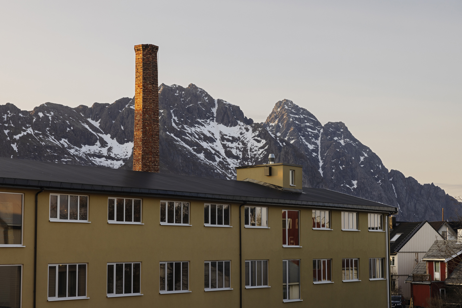 Foto av et langt gult hus med en høy murpipe, fjell i bakgrunnen.