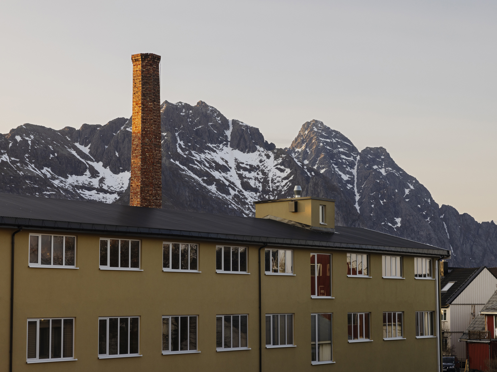 Foto av et langt gult hus med en høy murpipe, fjell i bakgrunnen.