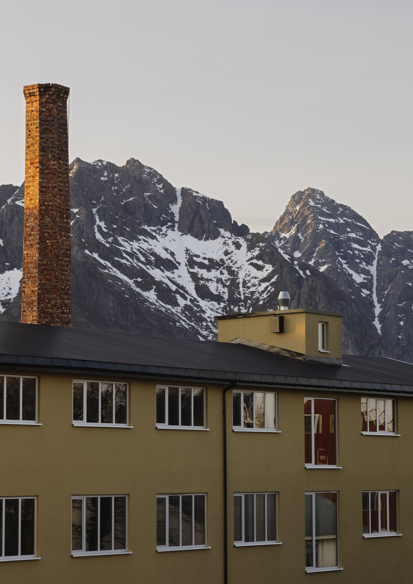 Foto av et langt gult hus med en høy murpipe, fjell i bakgrunnen.