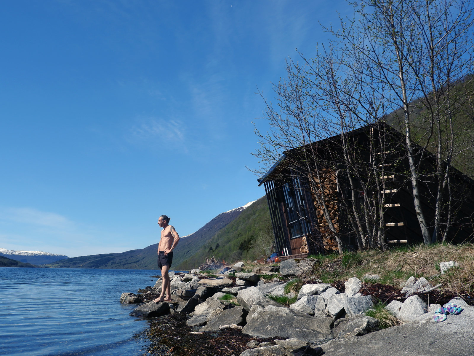 Foto av sauna som står i vannkanten, grønt landskap. Mann i shorts står og ser utover vannet. 