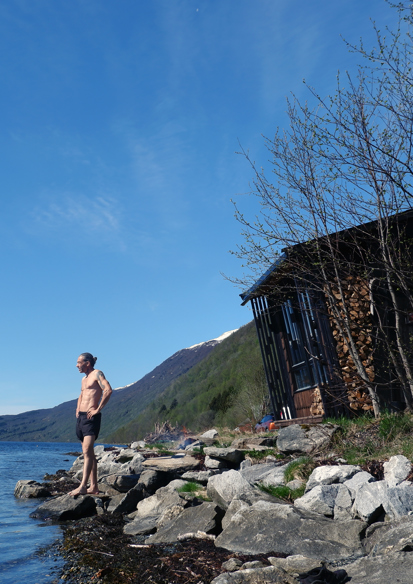 Foto av sauna som står i vannkanten, grønt landskap. Mann i shorts står og ser utover vannet. 