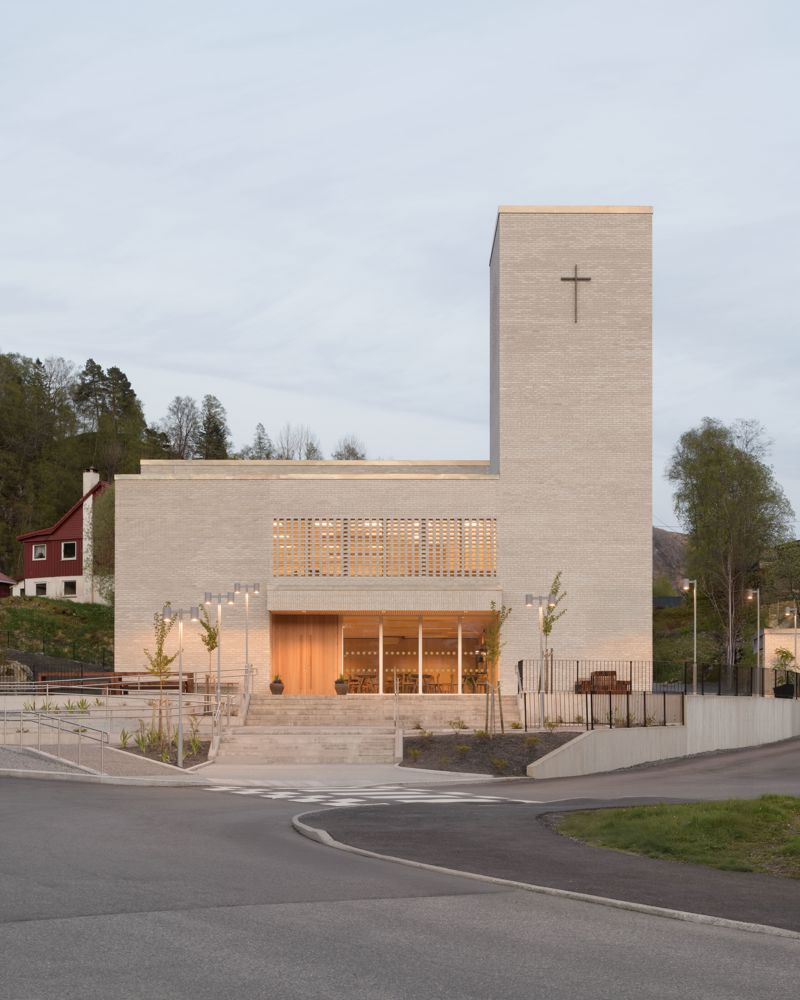 Foto av kirke i lys teglstein med varmt lys innenfra. Vei i forgrunnen.