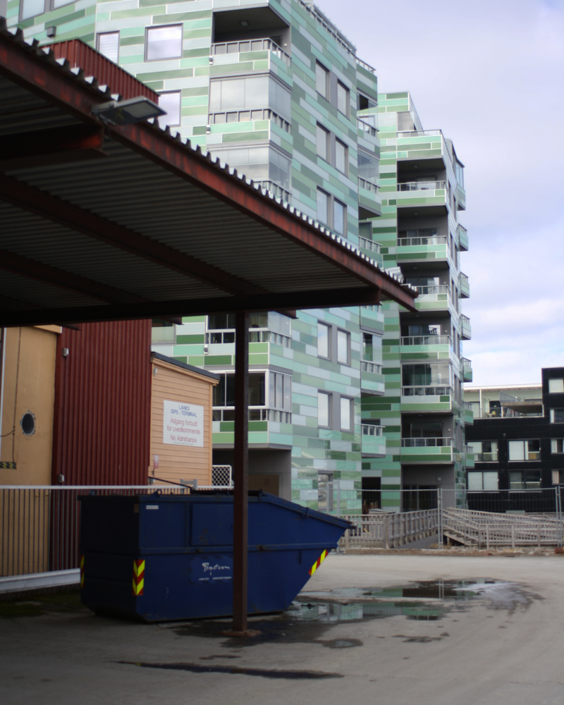 Foto av parkeringsplass med container, høyt leilighetsbygg bak i grønne toner.