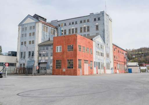 Foto av rødt og lyseblått industribygg, asfaltplass i forgrunnen. 