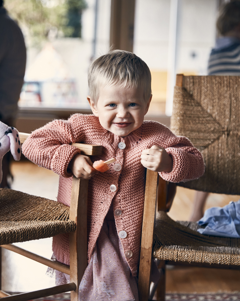 Foto av en liten jente i rosa kjole som står og smiler mellom to stoler.