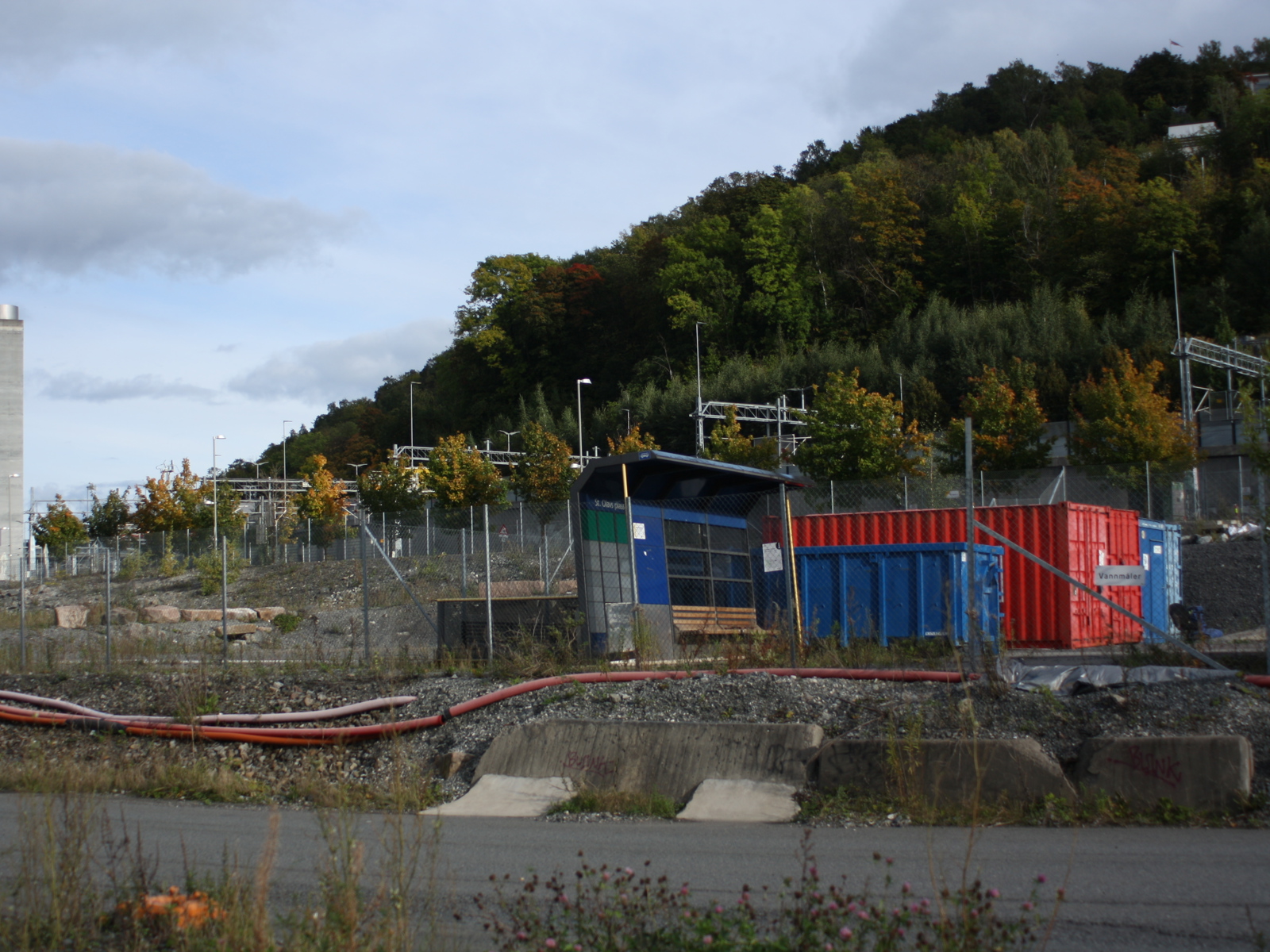 Foto av et rotete område ved siden av en vei, et gammelt busskur, containere med mer.