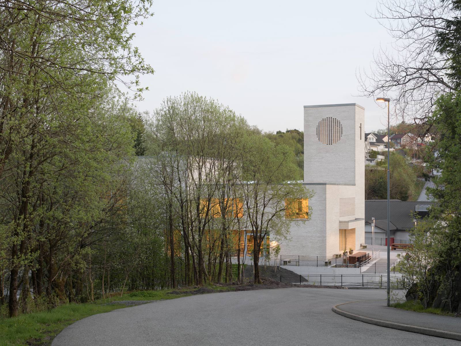 Foto av en vei som leder ned til en kirke i hvit teglstein, trær i forgrunnen.