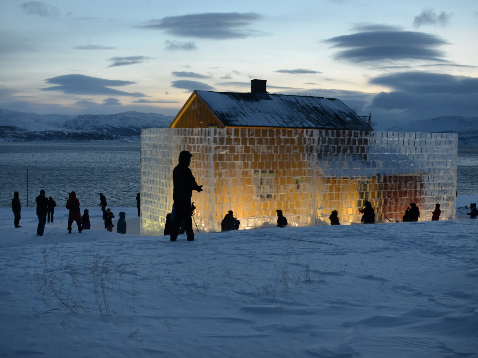 Foto av en is-skulptur ved et gult hus, skumring, mennesker som går rundt. 