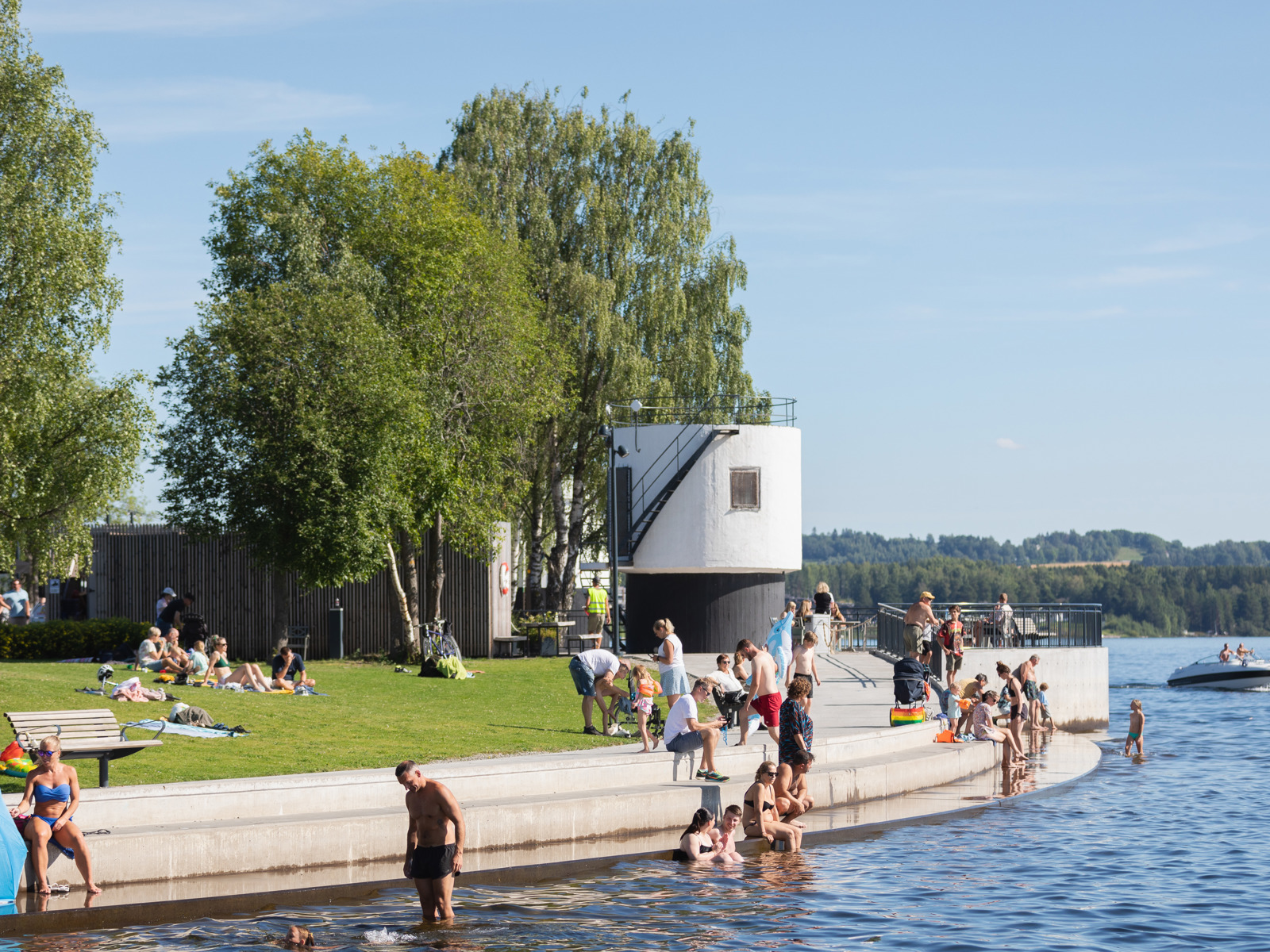 Foto av mennesker som bader og sitter i amfi langs vannkanten en fin sommerdag.