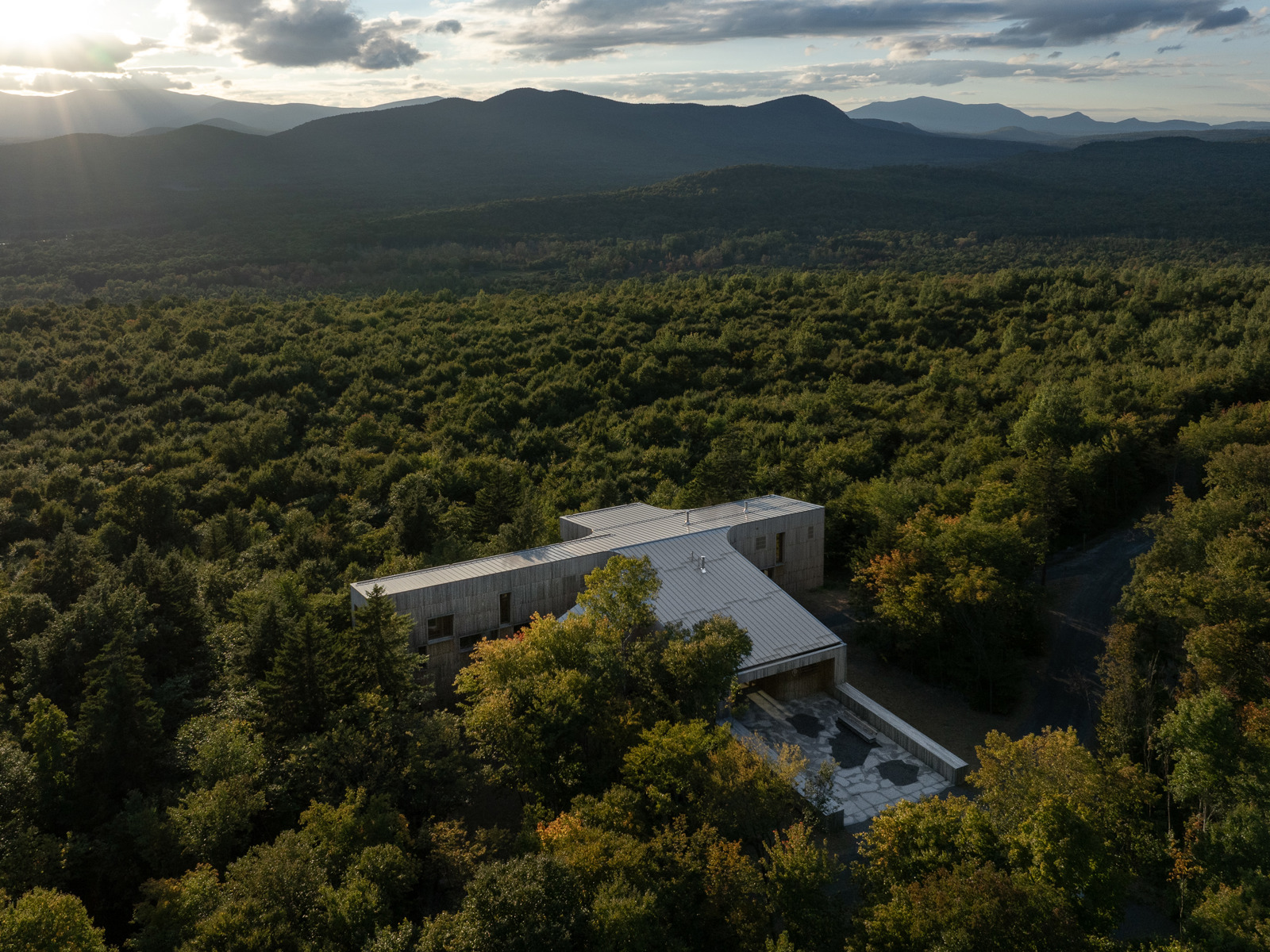Drone foto av et bygg som ligger helt alene i en stor skog, fjell i bakgrunnen.