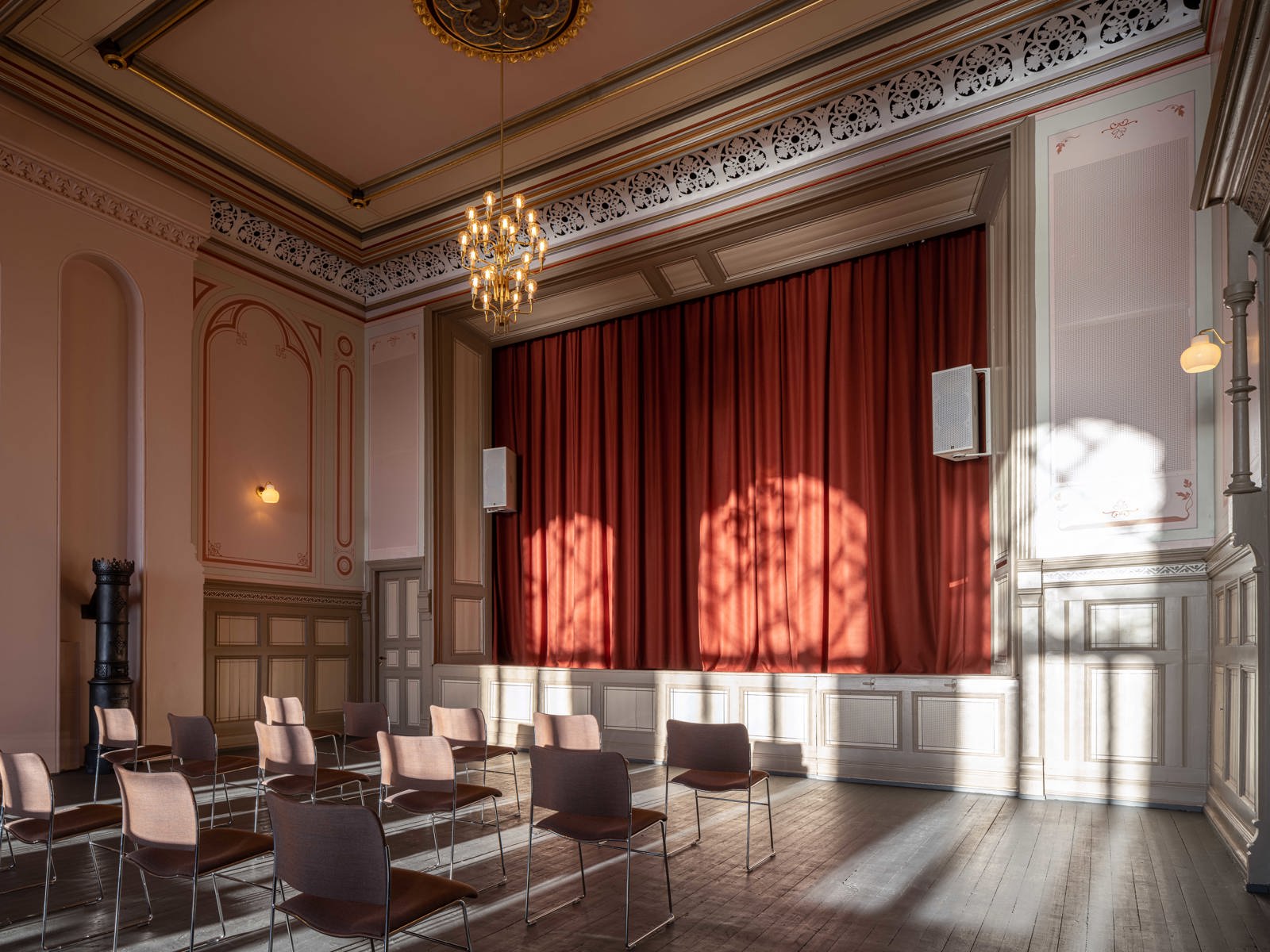 Interiør foto av festsalen i festiviteten, scene med gjendratte røde scenetepper, lysekrone og stoler plassert ut i salen.