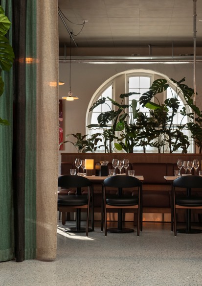 Interiørfoto av restaurant, pådekt bord, grønne planter, rundt vindu, gardiner.