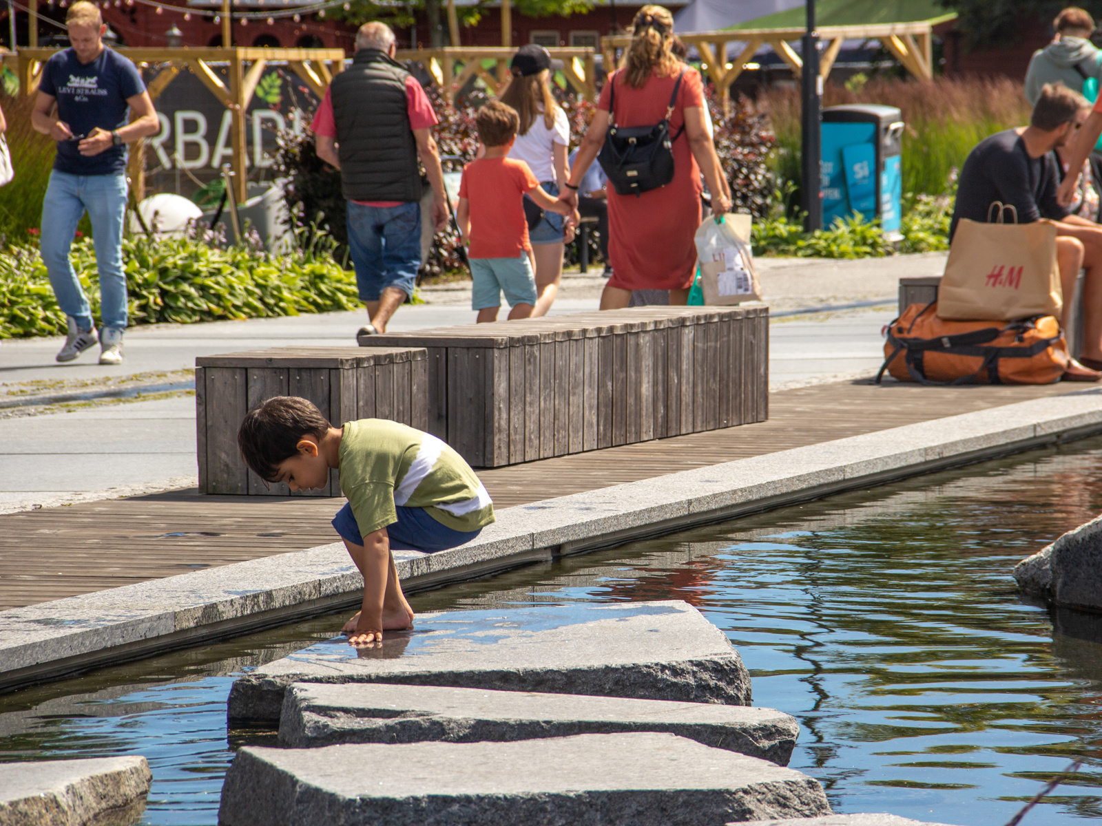 Gutt som står på en liten stein i en firkantet vanndam i en park, folk som går i bakgrunnen. Foto.