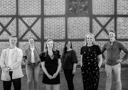 6 mennesker stående fremfor en vegg med eksponert bindingsverk, svart hvitt. Foto.