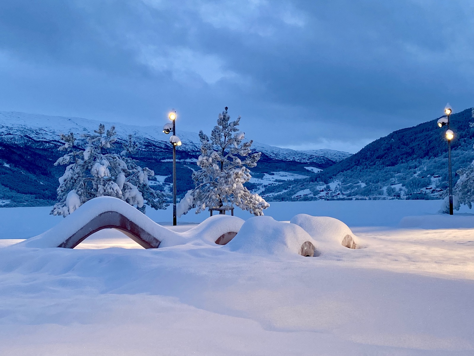 Foto av en skulptur benk i en snødekt park, fjell i bakgrunnen, kveldslys og gatelykter. 