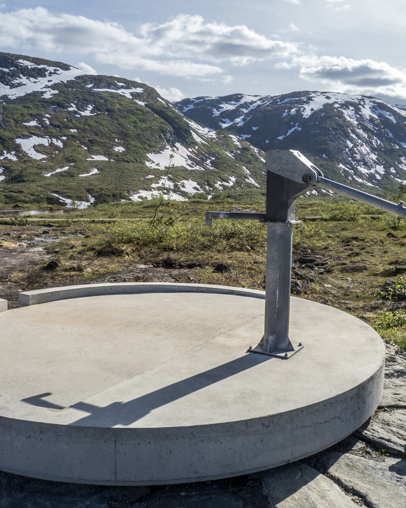 Vannpumpe på en rund betongplate, fjell med litt snø på i bakgrunnen. Foto.