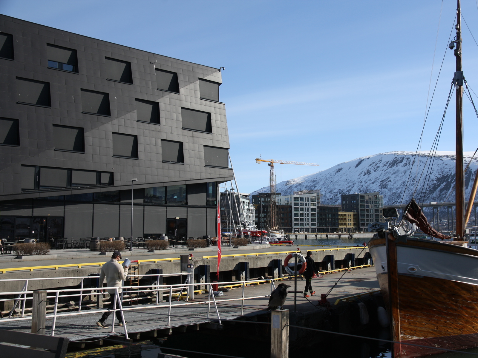 Foto av bryggekant, eldre trebåt i front, svart større bygg bak og fjell med snø lengst bak.