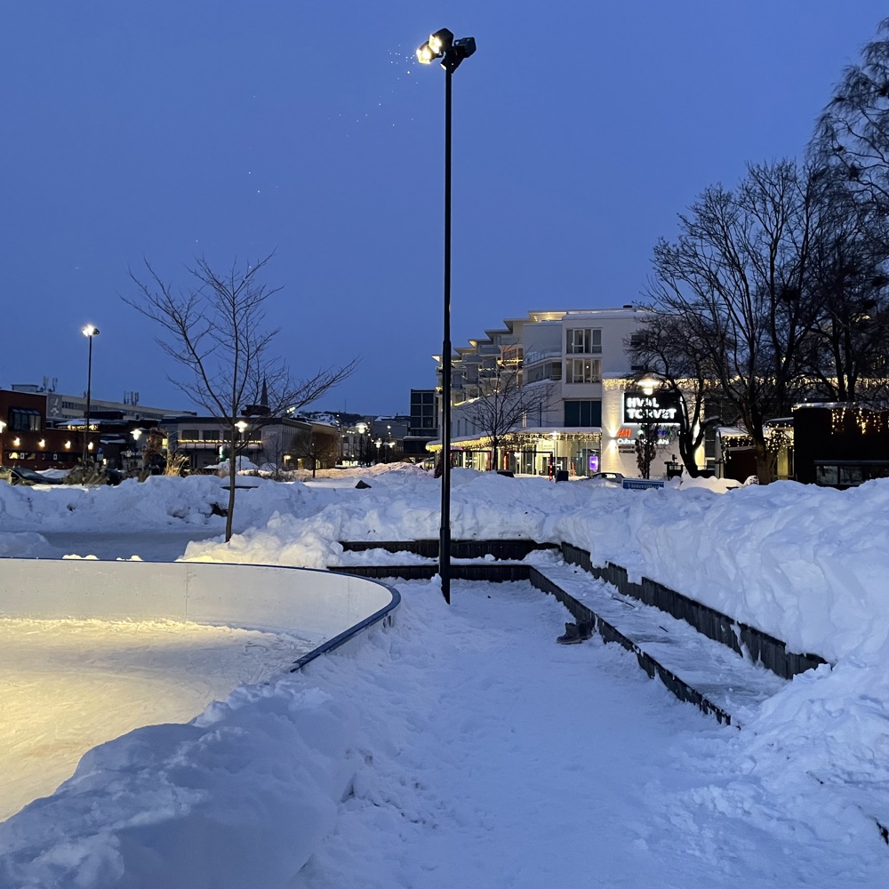 Skøytebane i snødekt park i blåtime, by i bakgrunnen. Foto.