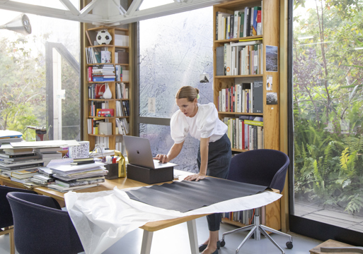 Foto av en kvinne på et kontor stående over en macbook, store vinduer ut til grønn natur.
