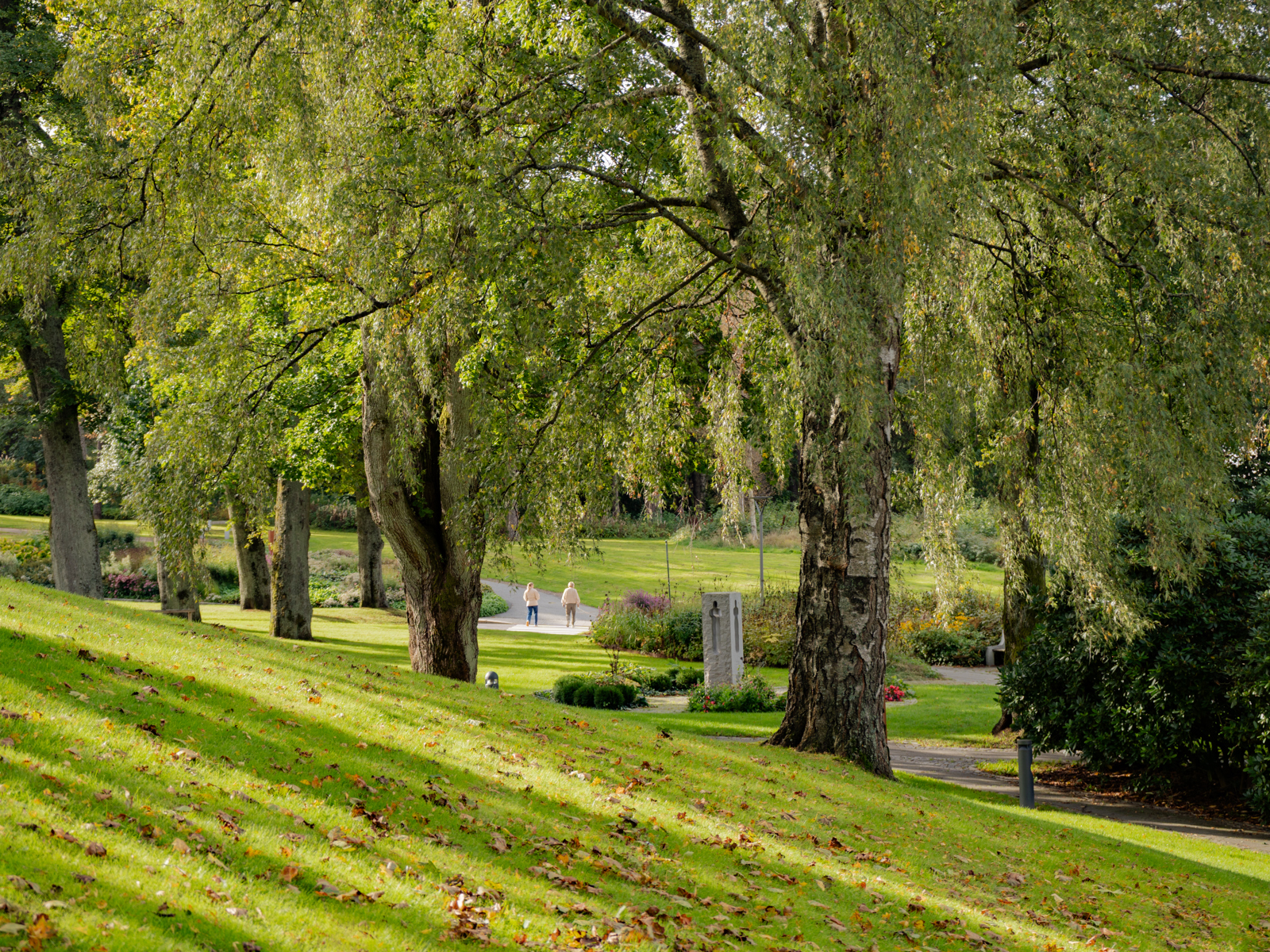 Foto av park med store trær, minnested og to mennesker som går på en grusvei.