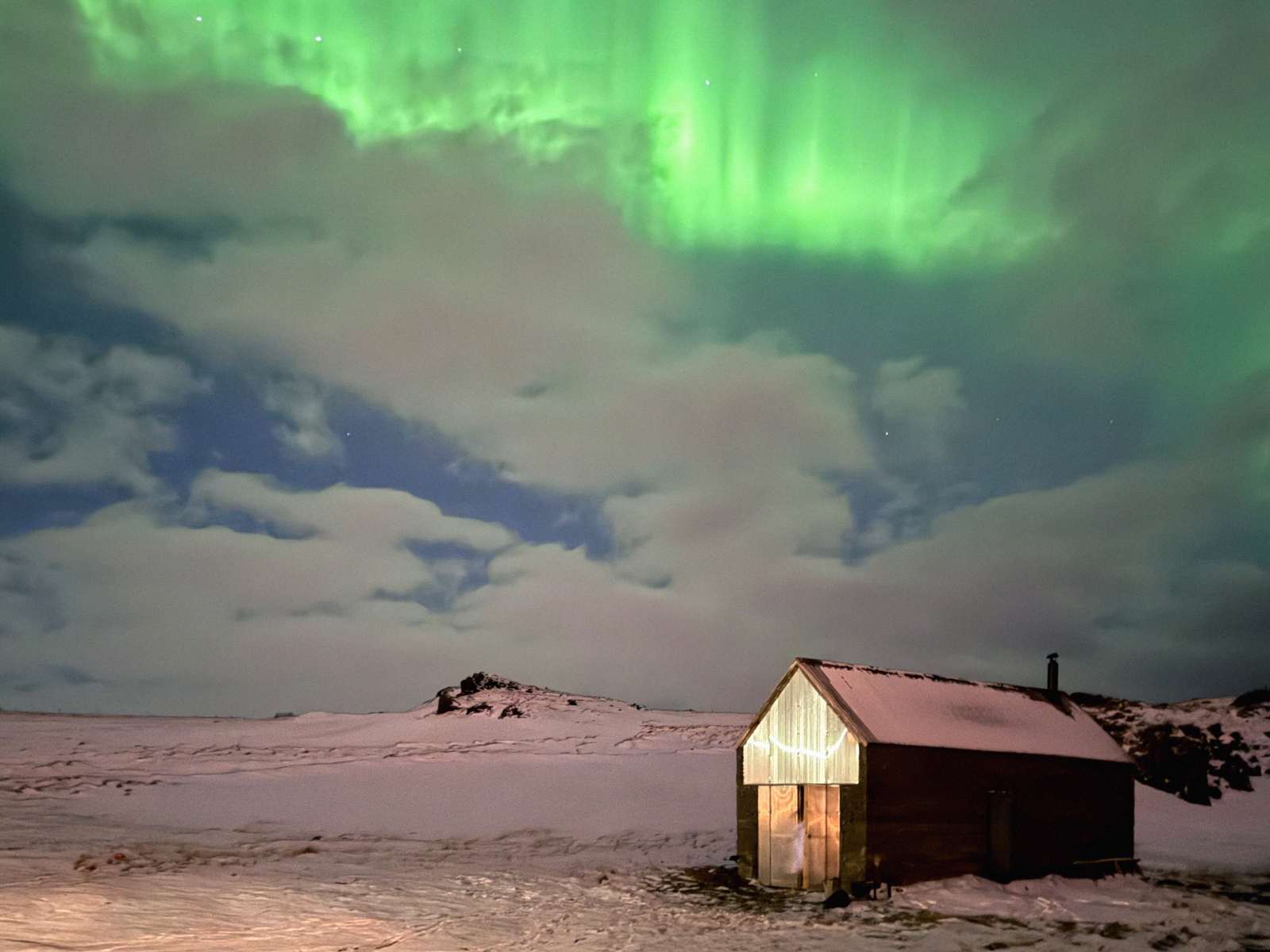 Foto av badstue som er opplyst innenfra  i snølandskap med nordlys over.